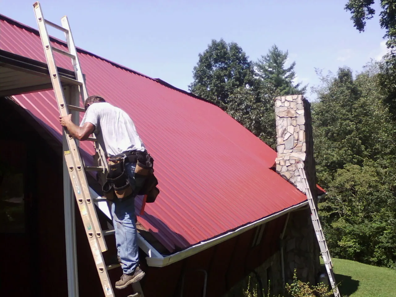 Roofer on a ladder beside a red metal roof and stone chimney — Soffit & Fascia Repair by Roofing Brothers in Mahopac